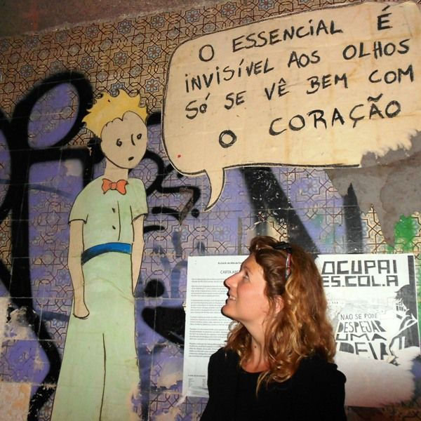 Antonia standing in front of wall, where there is a mural of Little Prince character, and text writen in Portugese.